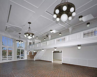 Interior of a former auto showroom showing the view from the mezzanine looking toward the upstairs balcony railing that extends the length of the room, with brick flooring, white painted woodwork, detailed celing with white globe chandeliers