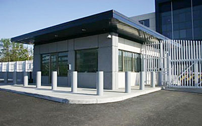 exterior of single story gatehouse surrounded by fixed bollards with white gate on right side