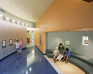 Colorful hallway with 2 children looking at pictures on the wall and a teacher and child reading in the niche built into the hall's wall