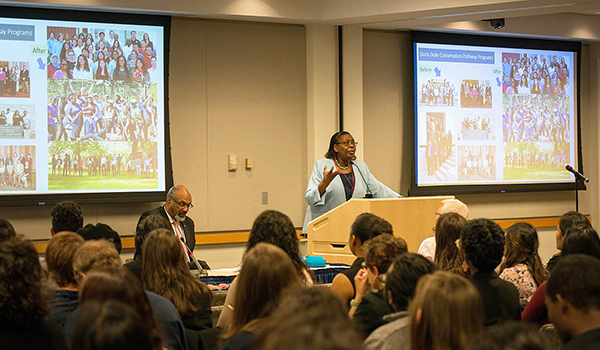 woman at a podium giving a lecture in front of a small audience with screen on the right and left behind her displaying visuals