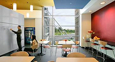 Small break room with a man writing on a wall sized white board, small tables and a feature window