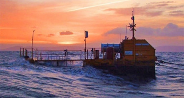 Photo of the OE Buoy, located in the ocean and designed around the oscillating water column principle. Waves surround the buoy and its turbine captures the wave energy, which the generator then converts into electrical power