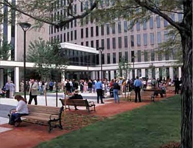 a plaza outside an office building where people are gathered standing along the sidewalks and sitting on the benches amongst the landscaping