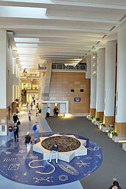 View of Winona States University's atrium which features a side wall displaying a mock-up of the shape, texture and geology of the bluffs along the Mississippi river. In the same space, a seating bench displays the original map of the constellations and is surrounded by key symbols of all disciplines within the building.